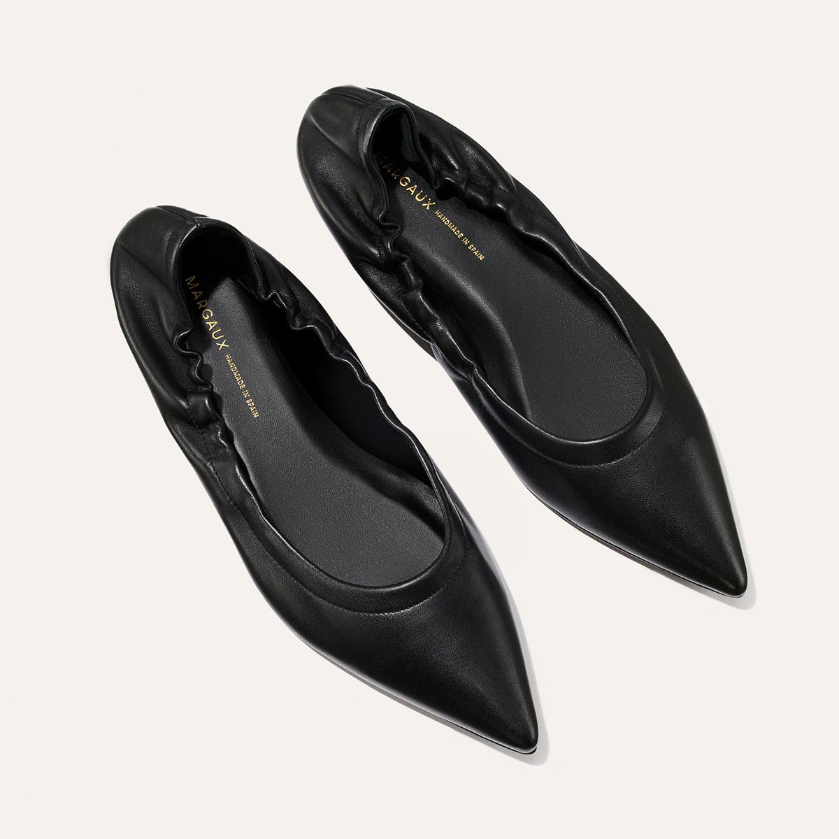 The Roma - Black Nappa by Margaux are elegant black pointed-toe ballet flats with ruched detailing, combining classic style and comfort, shown on a light background.