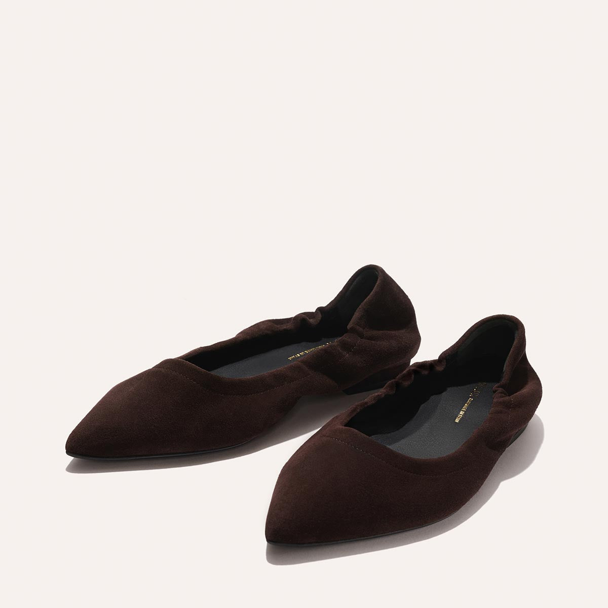 Two Espresso Suede "The Roma" ballet flats by Margaux with pointed toes and ruched detailing around the opening, shown on a light background. The insoles are black with gold Margaux branding.