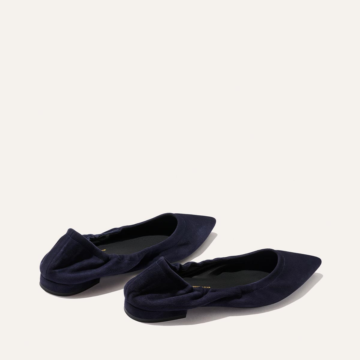 A pair of Margaux's The Roma ballet flats in indigo suede, featuring a pointed toe and ruched heel detailing, are displayed on a light background—bringing a sophisticated touch to any ensemble.