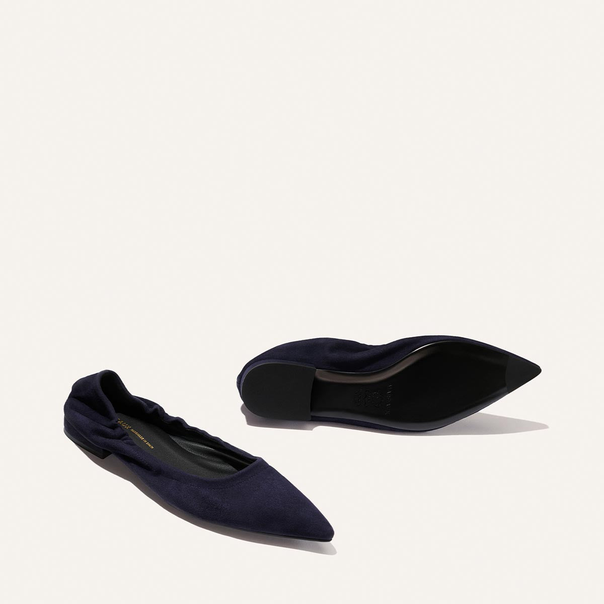 A pair of The Roma - Indigo Suede ballet flats by Margaux, featuring a navy blue pointed toe design—one shoe displayed upright and the other revealing its black sole—set against a light background. These elegant flats blend classic sophistication with contemporary versatility.