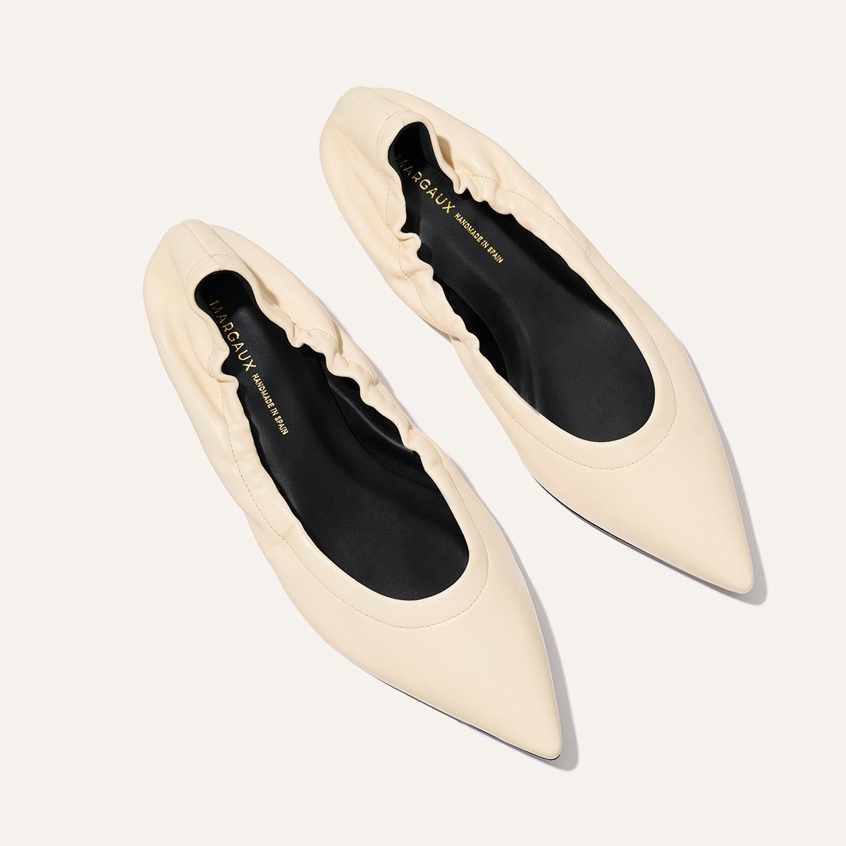 A pair of Margaux's The Roma - Ivoire Nappa cream-colored pointed-toe ballet flats with ruched edges, shown on a light background. They feature black insoles with gold branding printed near the heel.