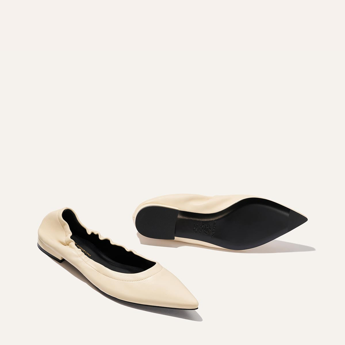 The Roma - Ivoire Nappa by Margaux: cream-colored pointed-toe ballet flats with black soles, one upright and one on its side, shown on a plain light background.