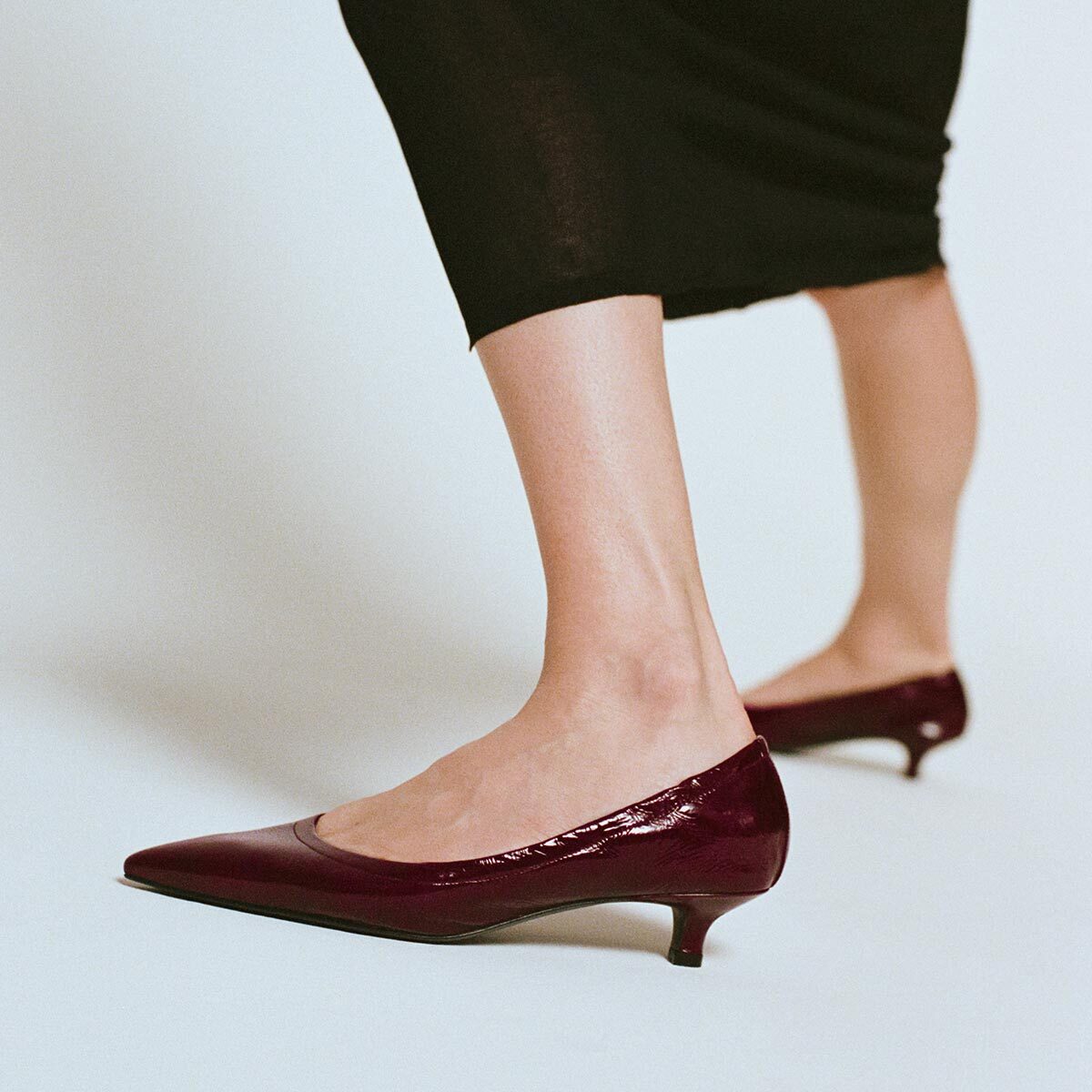 A woman wearing Margaux’s The Roma 40 - Merlot Crinkle Patent pumps with a walkable heel and a black skirt, photographed from the knees down against a light gray background.