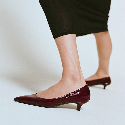 A woman wearing Margaux’s The Roma 40 - Merlot Crinkle Patent pumps with a walkable heel and a black skirt, photographed from the knees down against a light gray background.
