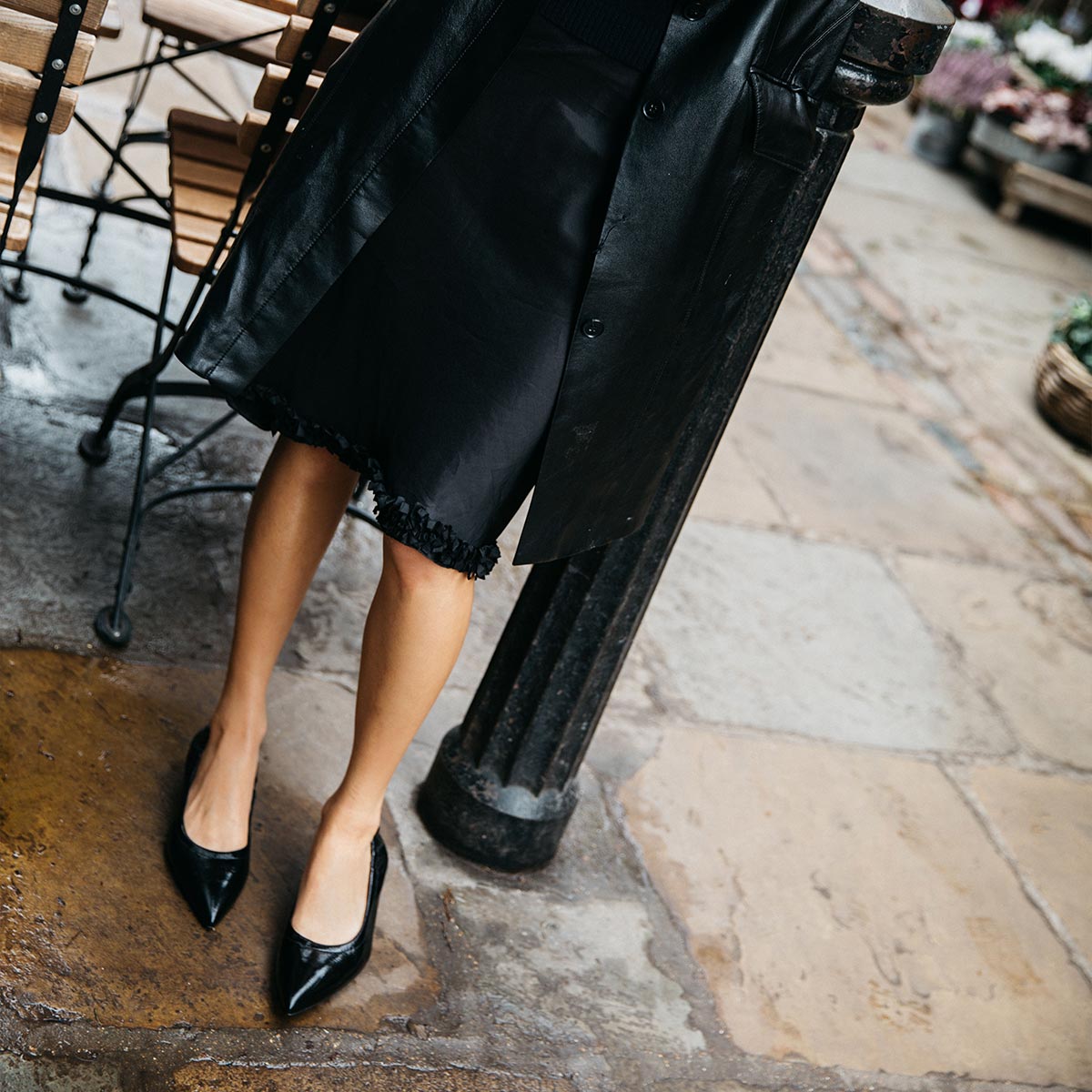 A person stands on a stone sidewalk beside a metal post and wooden chair, wearing Margaux's The Roma 40 - Black Crinkle Patent shoes, a black coat, and a black lace-trimmed skirt.