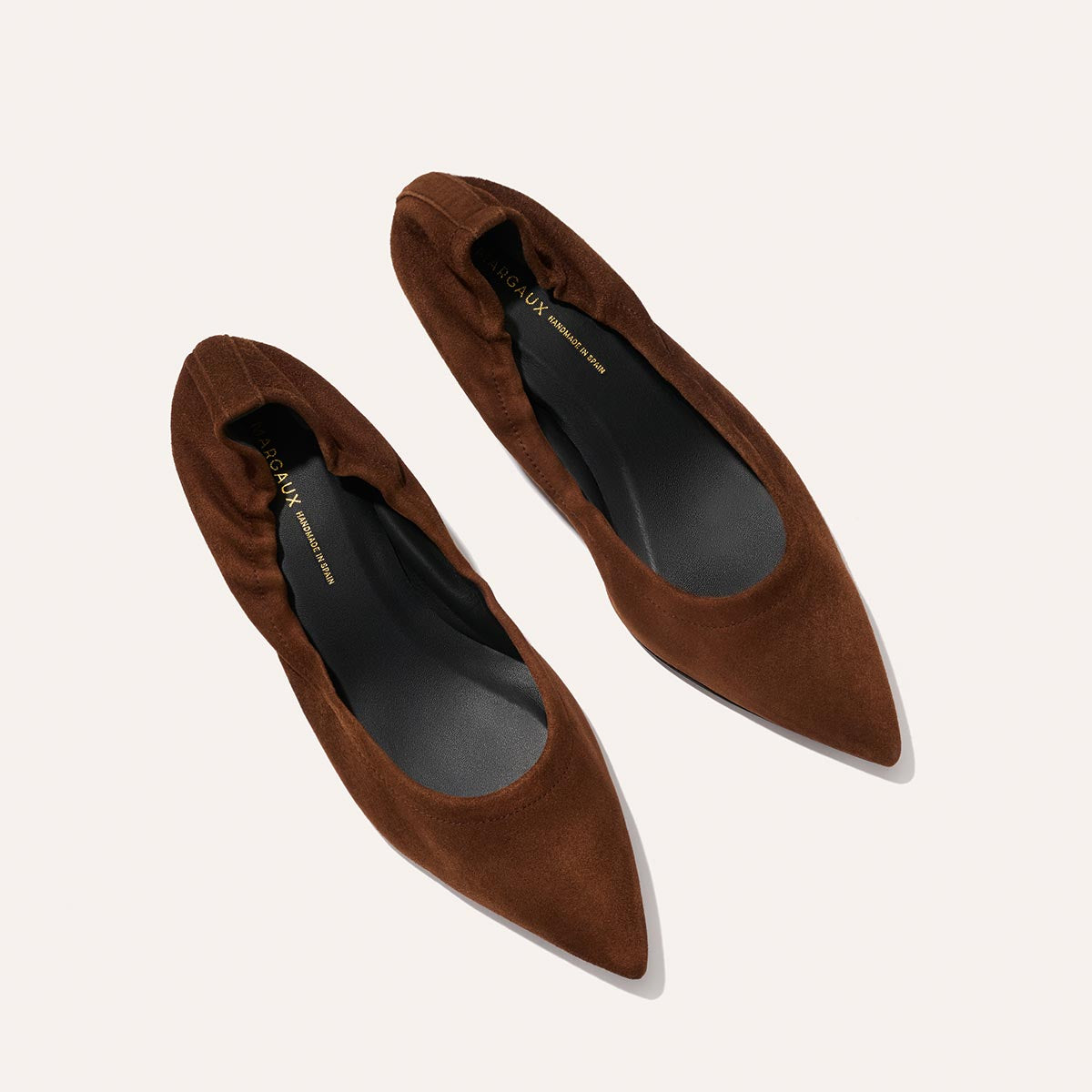 A pair of The Roma 40 - Ganache Suede pointed-toe flats by Margaux, shown from above with black insoles on a light background, offer classic elegance and a comfortable, walkable heel.