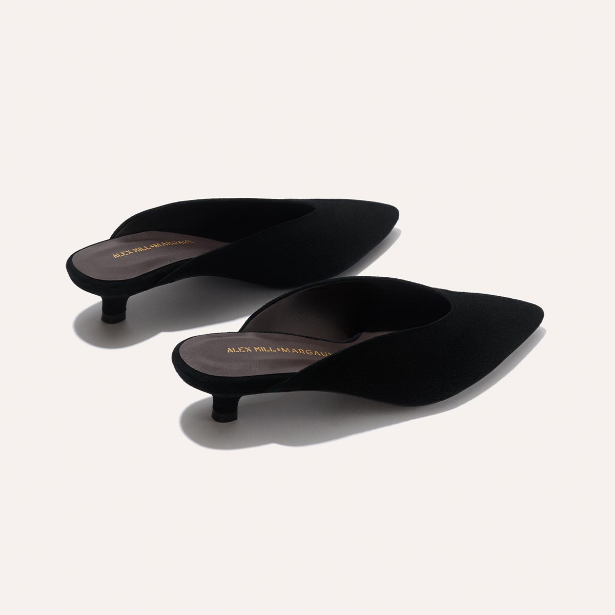 The Romy - Black Suede kitten mule heels by Margaux, from the Alex Mill x Margaux collection, feature pointed toes, black slip-on design, and gold branding on the insoles, displayed on a neutral background.