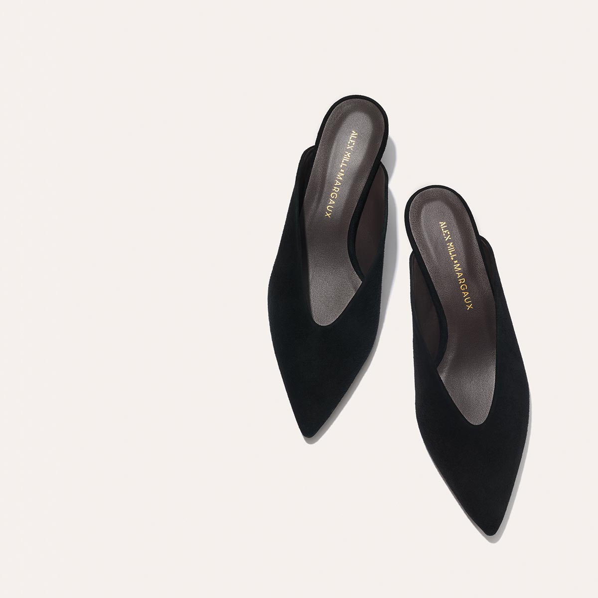 The Alex Mill x Margaux: The Romy - Black Suede features black pointed-toe kitten mules with a low heel, ideal for parties. "Margaux" is printed in gold on the insole, shown against a light beige background.