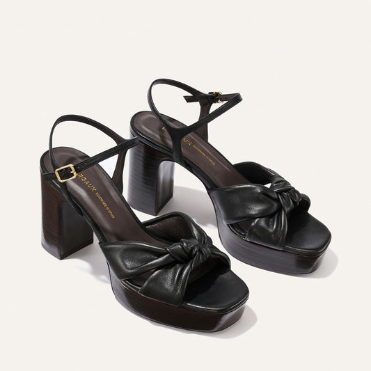 The Sabine Platform by Margaux in Black Glove Nappa features chunky high heels, a knotted front strap, and slim ankle straps with small buckles, showcased against a plain light background.