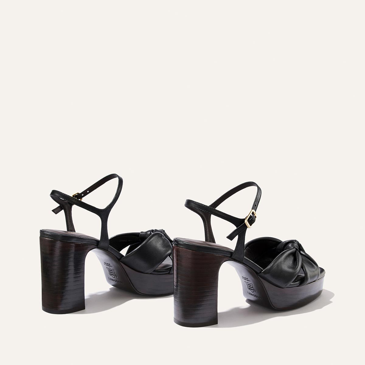 A pair of Margaux’s The Sabine Platform sandals in Black Glove Nappa, featuring ankle straps and chunky brown wooden heels, are shown against a plain light background, viewed from the back.