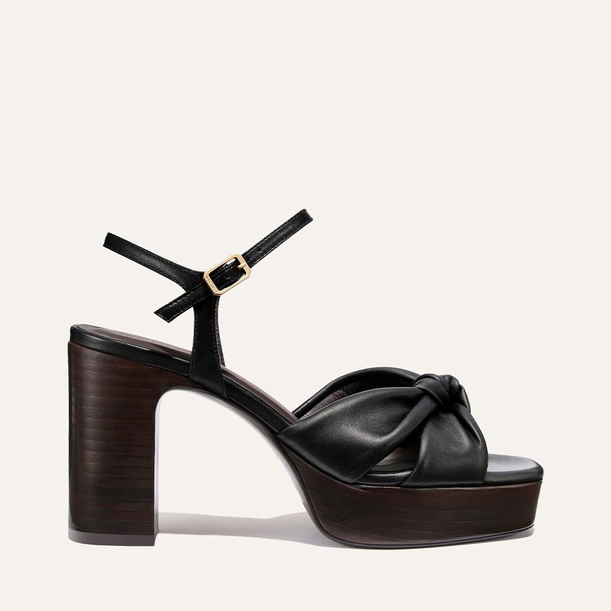 The Sabine Platform by Margaux in Black Glove Nappa features a thick wooden platform, a knotted leather front strap, and an ankle strap with a gold buckle, set against a plain light background.
