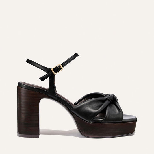 The Sabine Platform by Margaux in Black Glove Nappa features a thick wooden platform, a knotted leather front strap, and an ankle strap with a gold buckle, set against a plain light background.