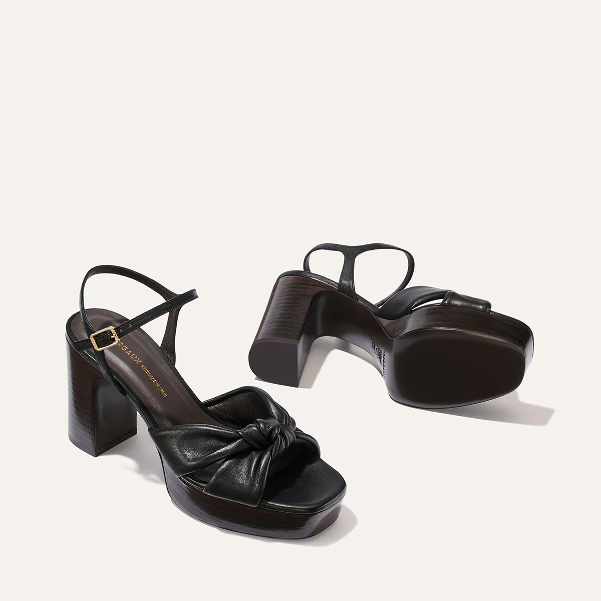 Introducing The Sabine Platform by Margaux in Black Glove Nappa: a pair of black, high-heeled sandals featuring ankle straps and a knotted leather detail at the toe, shown from side and bottom angles on a light background.