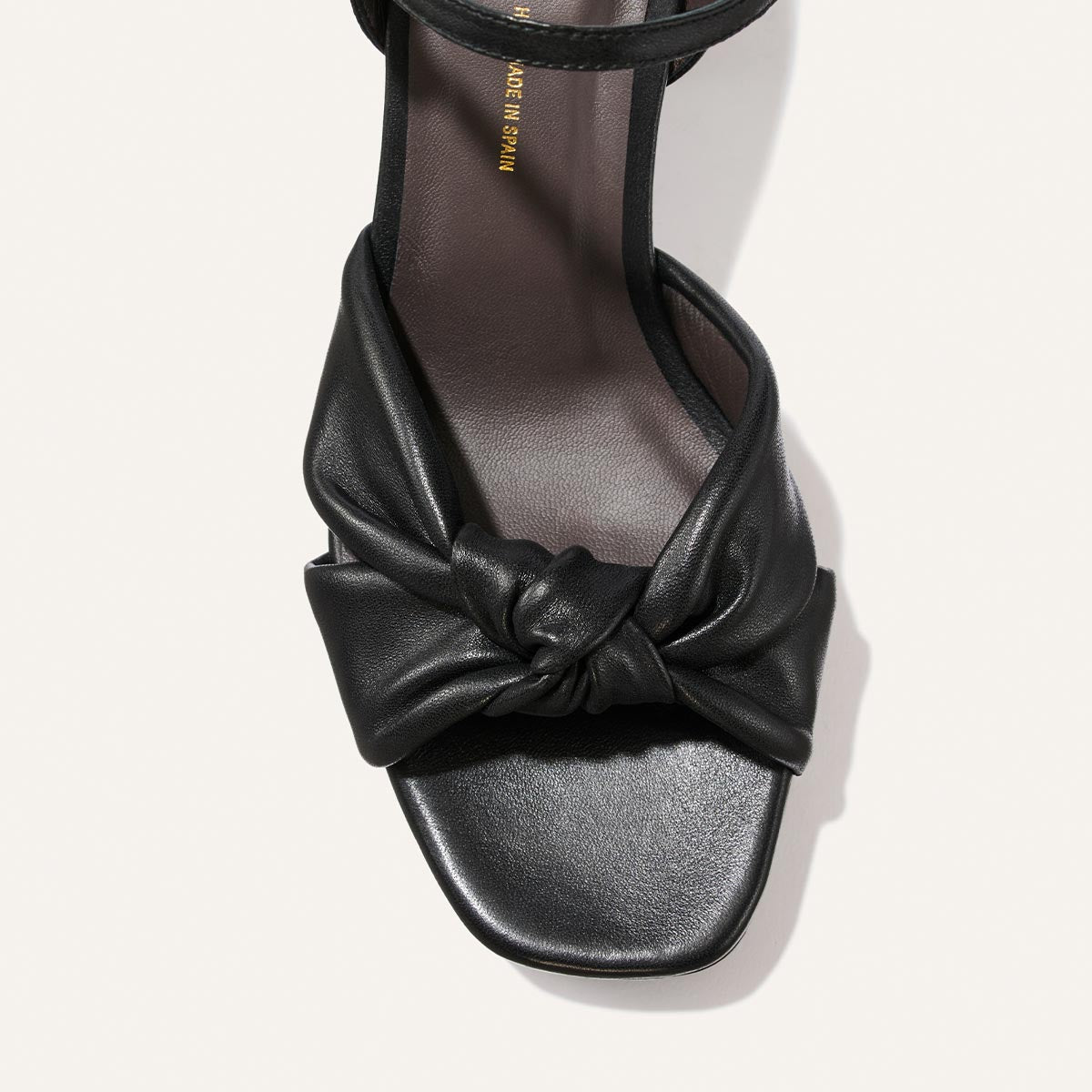The Sabine Platform by Margaux features a black glove nappa leather construction with a twisted knot detail on the front strap, an open toe, and a squared front sole. The sandal has a dark footbed made of smooth leather, as shown in the top-down image.