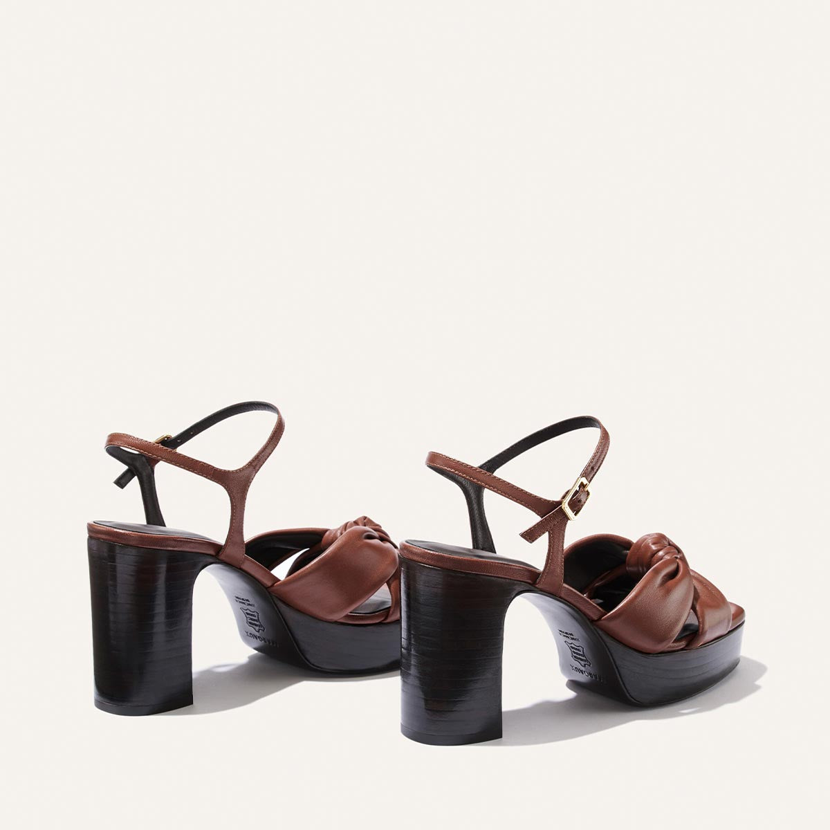 The Sabine Platform by Margaux in Brandy Glove Nappa features brown high heels with thick, dark wooden platforms, ankle straps, and knotted leather detailing on the front, displayed on a plain white background.