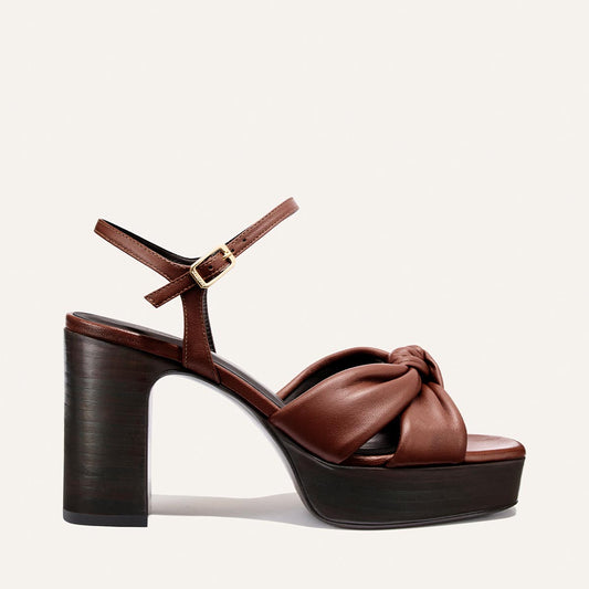 The Sabine Platform by Margaux in Brandy Glove Nappa—a brown leather sandal with a thick high heel, a knotted front strap, and an adjustable ankle strap with a gold buckle—is displayed on a plain light background.