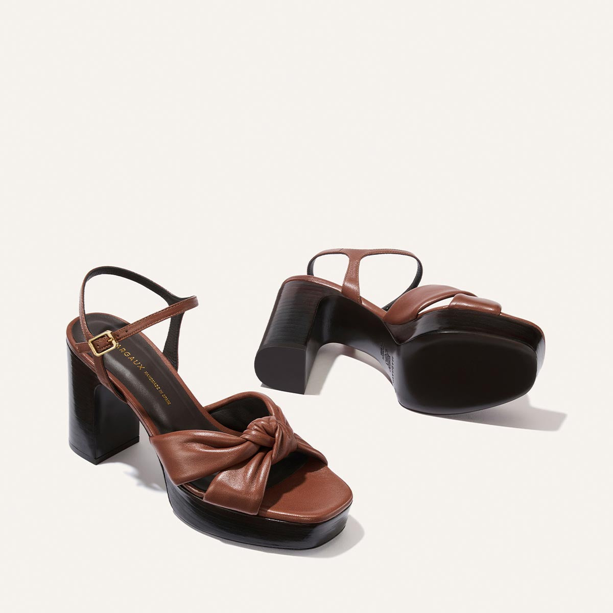 A pair of The Sabine Platform sandals in Brandy Glove Nappa by Margaux, featuring brown knotted straps and chunky black heels, are displayed on a light background—one upright and the other on its side to reveal the sole.