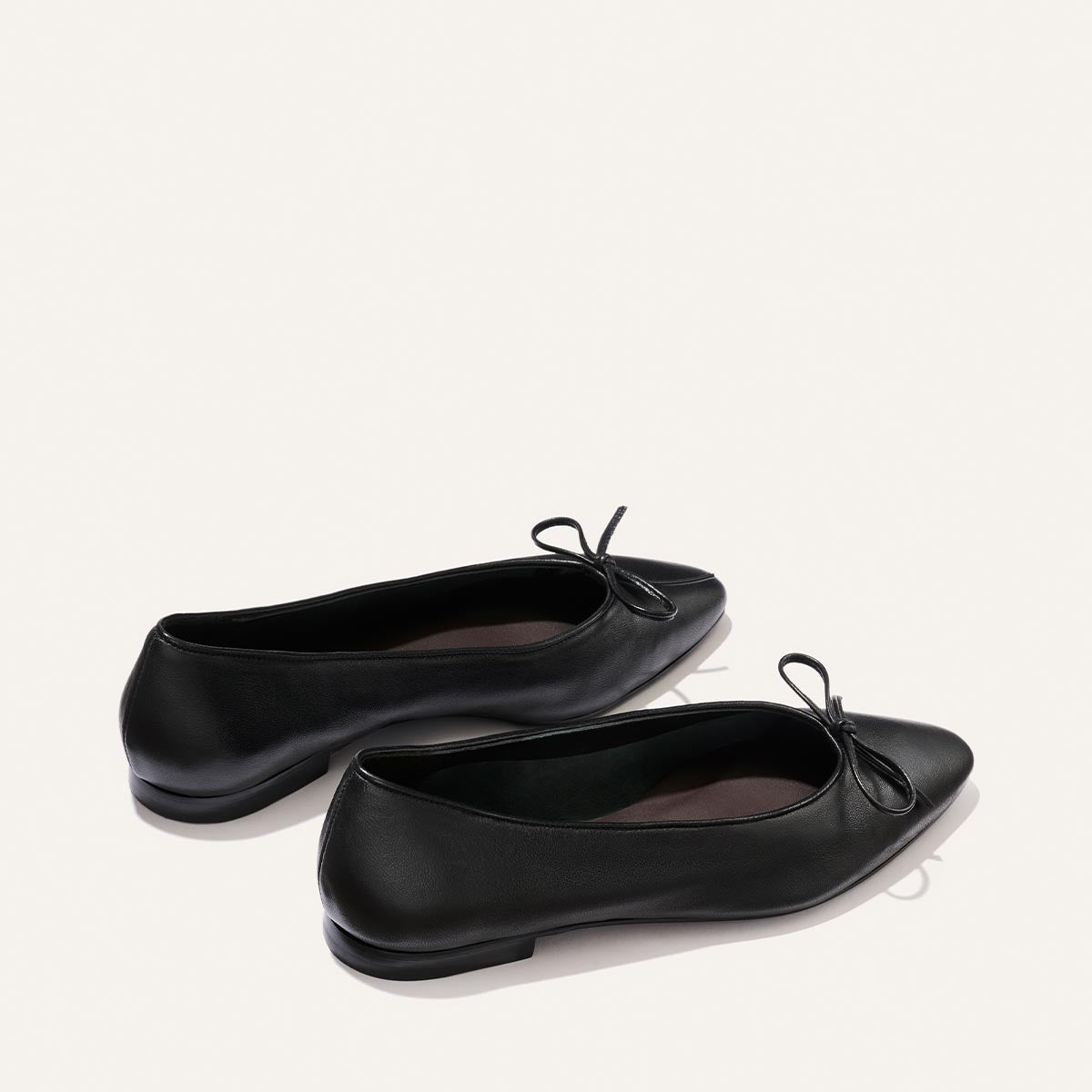 A pair of The Salamanca - Black Nappa ballet flats by Margaux, featuring small bows on the toes, displayed on a light background.