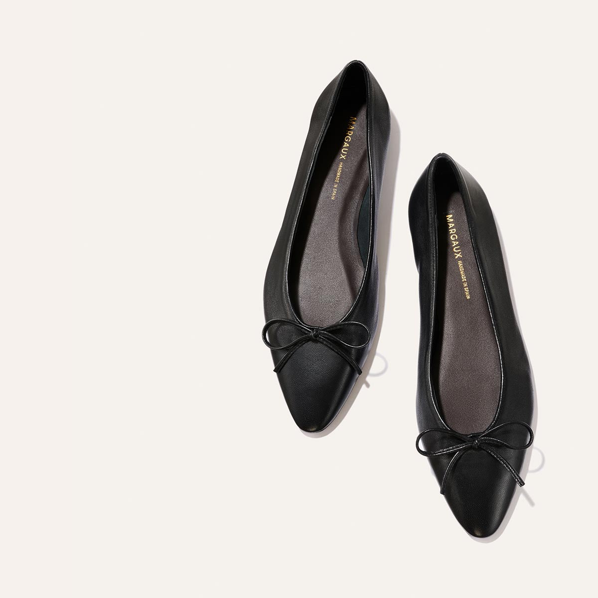 A pair of Margaux's The Salamanca - Black Nappa pointed-toe ballet flats, featuring small bows on the front, is displayed on a light-colored background.