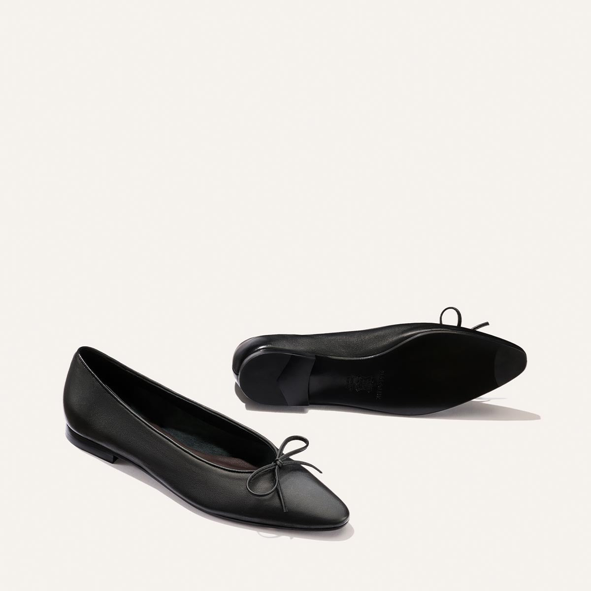 A pair of Margaux’s The Salamanca - Black Nappa ballet flats with pointed toes and small bow accents, displayed on a light background with one shoe upright and the other lying on its side.