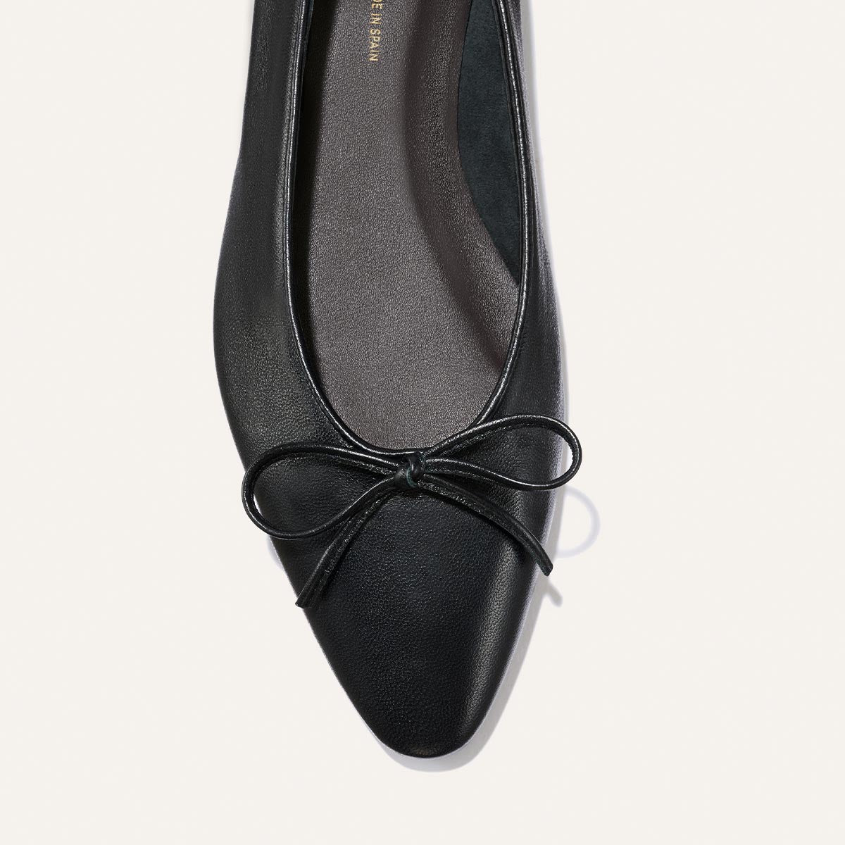 A close-up of The Salamanca - Black Nappa flat by Margaux, featuring a pointed toe and a small bow on the front, set against a light background.