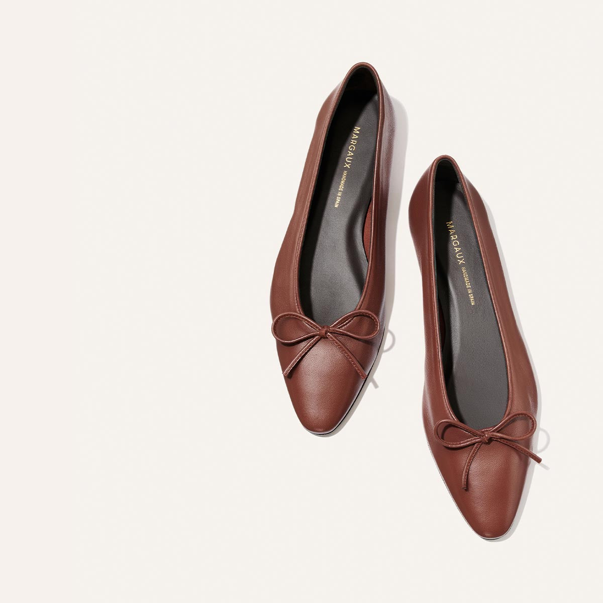 A pair of “The Salamanca - Brandy Nappa” pointed toe ballet flats by Margaux, crafted from rich brown leather and adorned with small bows on the front, elegantly displayed on a light background. These sophisticated shoes blend classic charm with modern style, making them a perfect addition to any wardrobe.