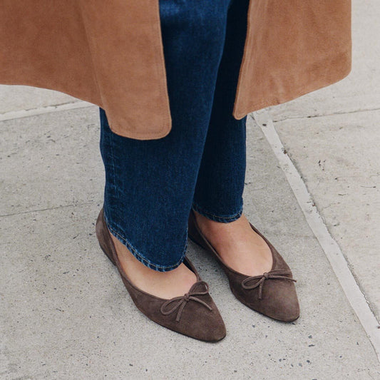 A person wearing dark blue jeans, a tan coat, and Margaux’s Alison Roman x Margaux: The Salamanca ballet flats in English Brown Suede—these unlined flats add a touch of elegance—stands on a gray sidewalk.