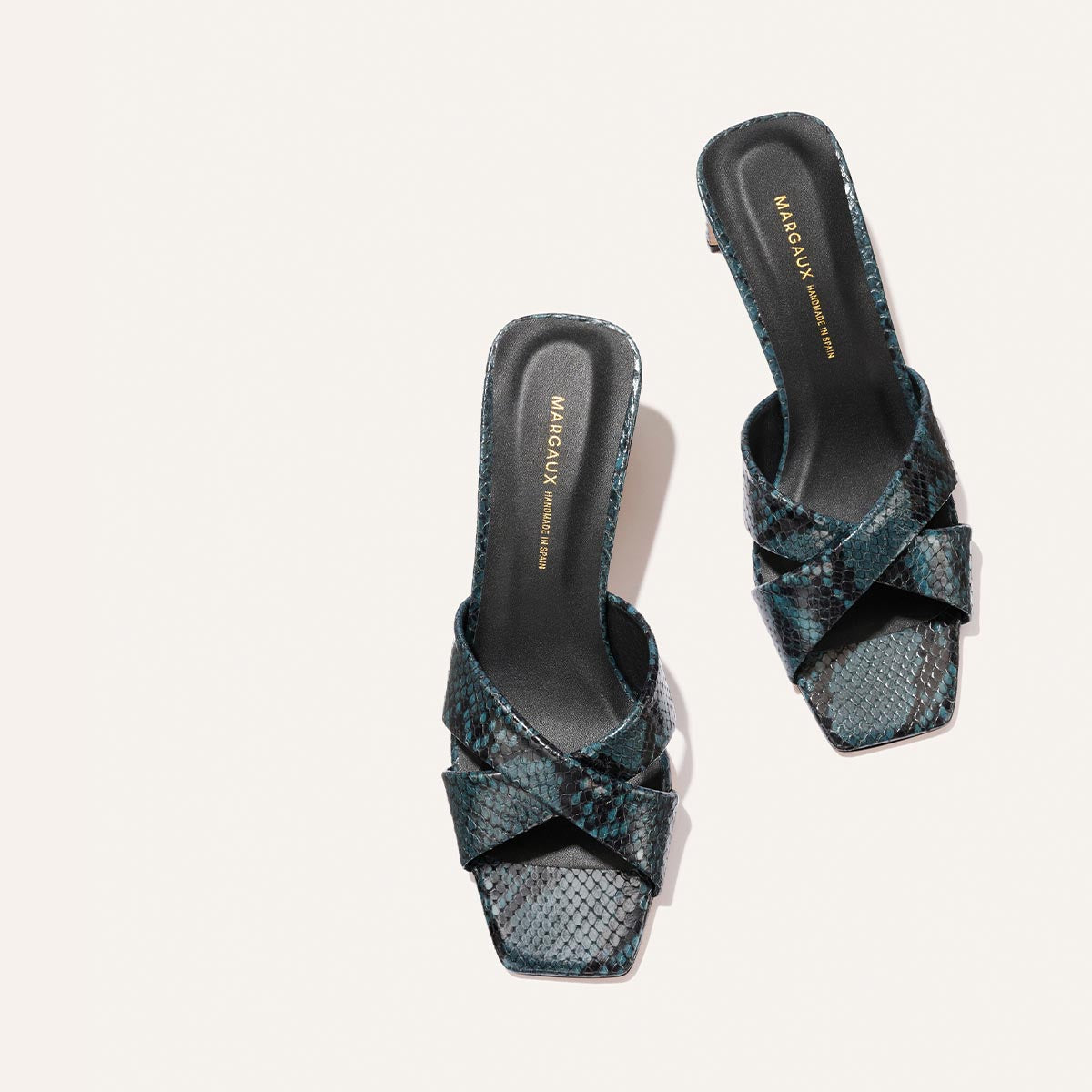 The Selene 50 - Aqua Python Embossed sandals by Margaux feature crisscross straps, a dark blue and black snakeskin pattern, square toes, and “Margaux New York” insoles, shown against a light background.