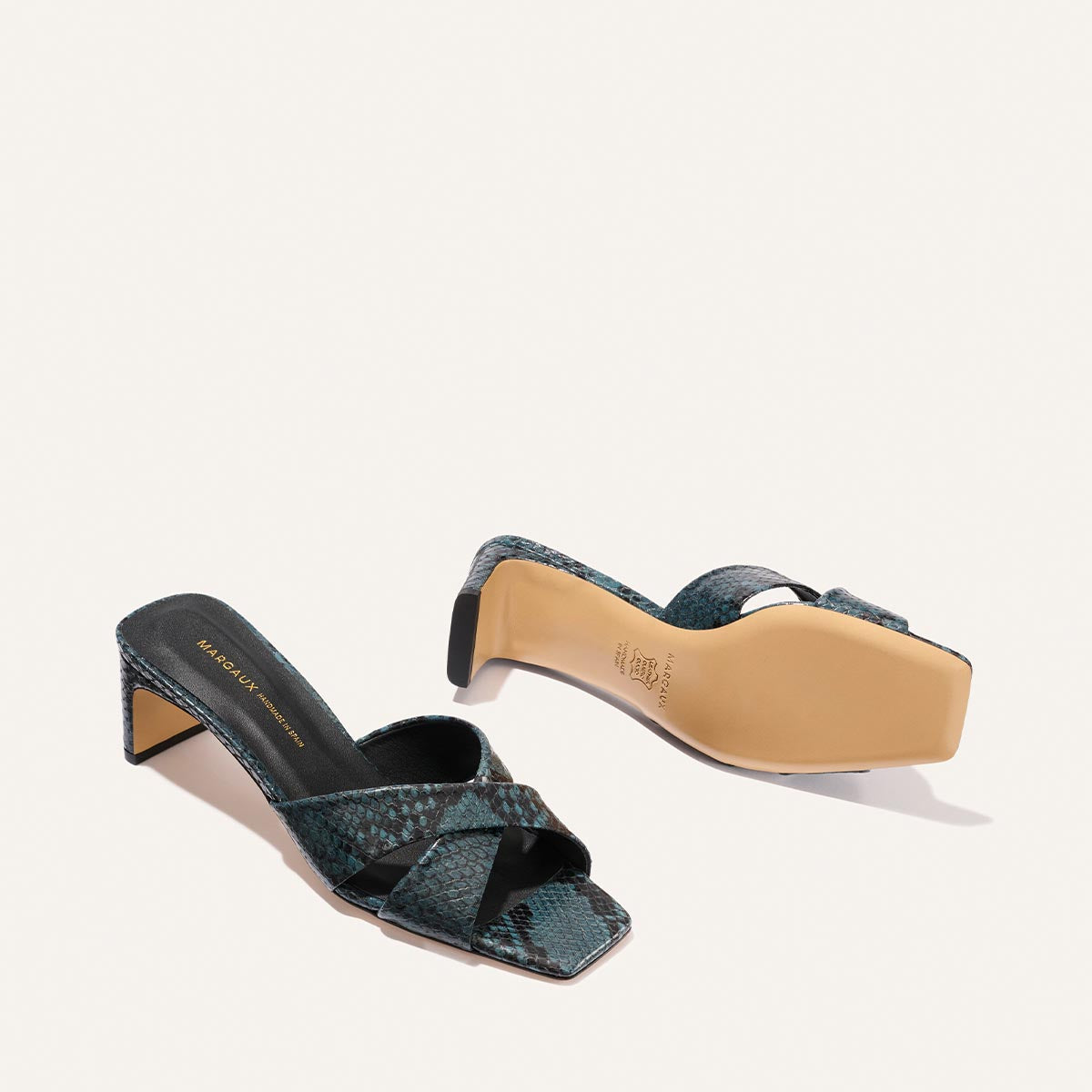 The Margaux Selene 50 - Aqua Python Embossed features heeled sandals with a blue-green snakeskin pattern, square toe, and crisscross straps. One shoe stands upright; the other lies on its side, revealing a beige sole and low block heel.