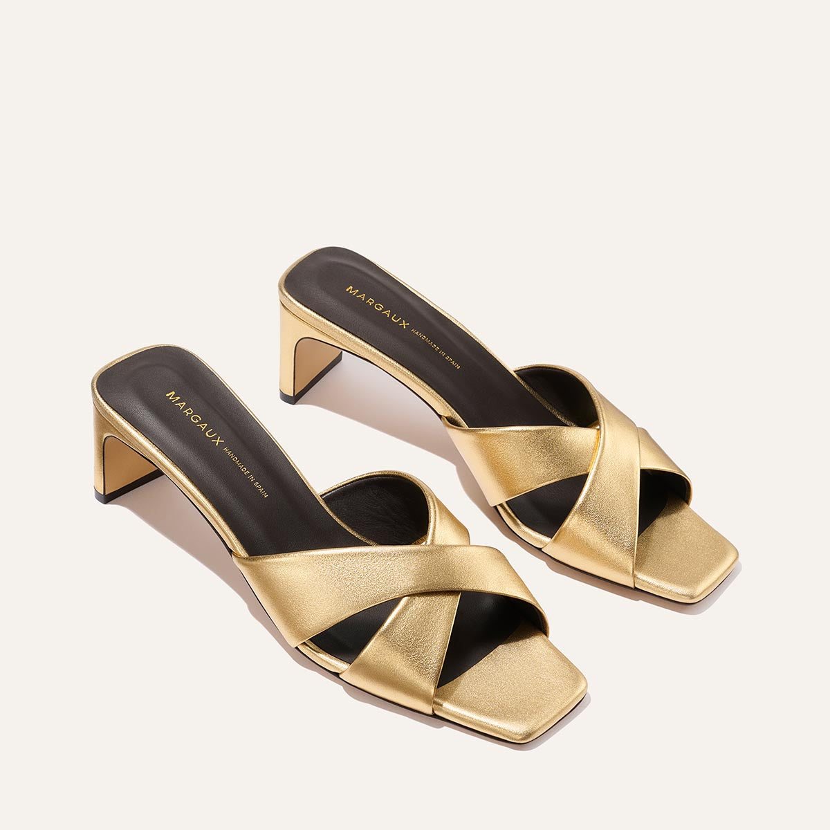 The Margaux Selene 50 - Gold Nappa features gold leather crisscross straps, an open square toe, black insoles with gold branding, and a chic block heel—elegantly displayed on a light background.