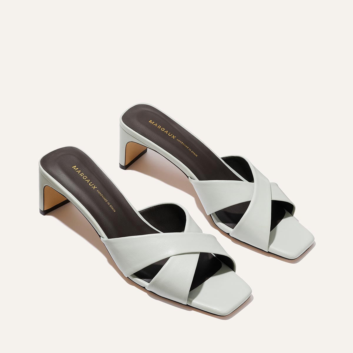 The Selene 50 - Sky Nappa by Margaux features white block heels, crisscross leather straps, square toes, and chunky heels, beautifully displayed on a light background.