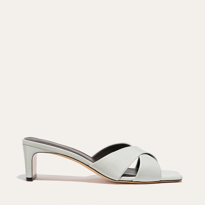 The Selene 50 - Sky Nappa by Margaux is a light gray heeled sandal with a low square block heel and open toe. It features two wide, crisscrossed leather straps over the foot and is displayed on a plain white background.