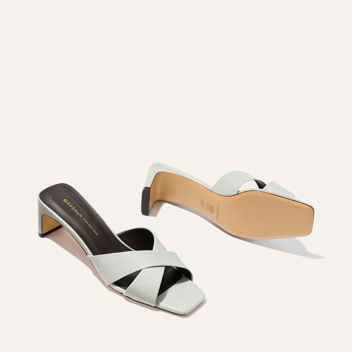 Two Selene 50 - Sky Nappa heeled sandals by Margaux in light gray with leather criss-cross straps and square toes are shown; one stands upright, the other lies on its side to reveal the sole.