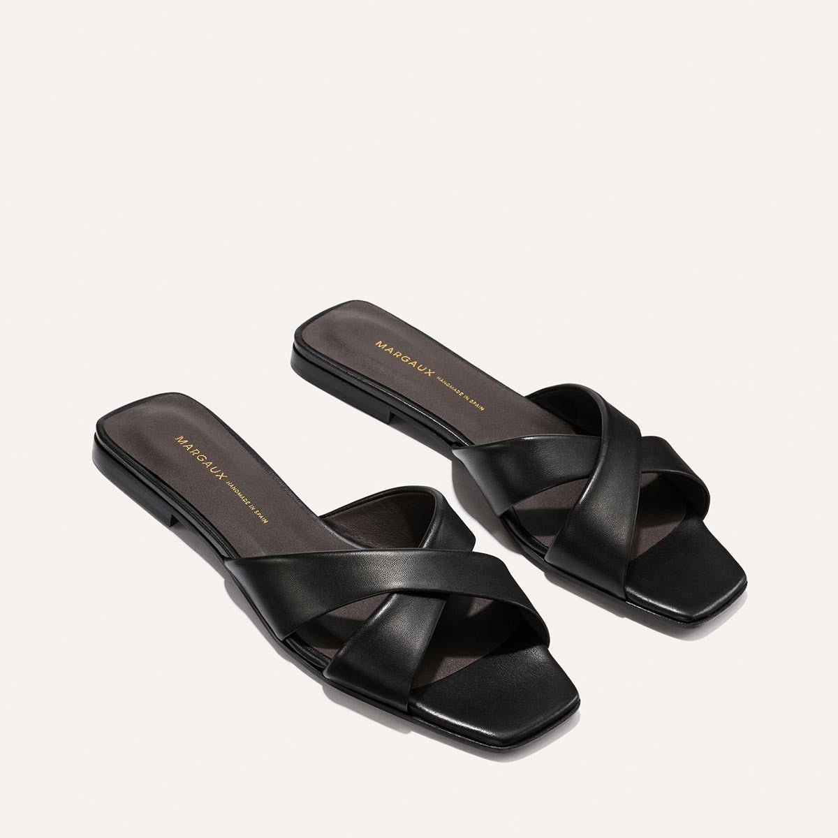 The Selene Flat Sandal by Margaux in Black Nappa features crisscross leather straps, square toes, and dark insoles with gold branding, displayed on a light background.