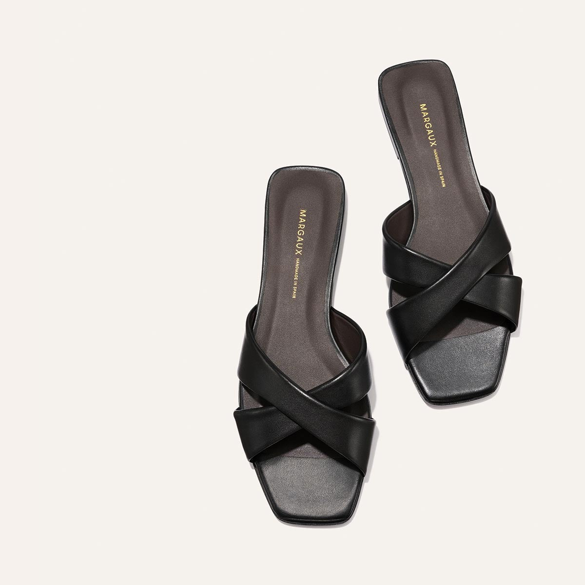 The Selene Flat Sandal by Margaux in Black Nappa features crisscross straps, a square toe, and a slip-on design. Displayed on a light background, the sandal has a dark insole with gold branded lettering.