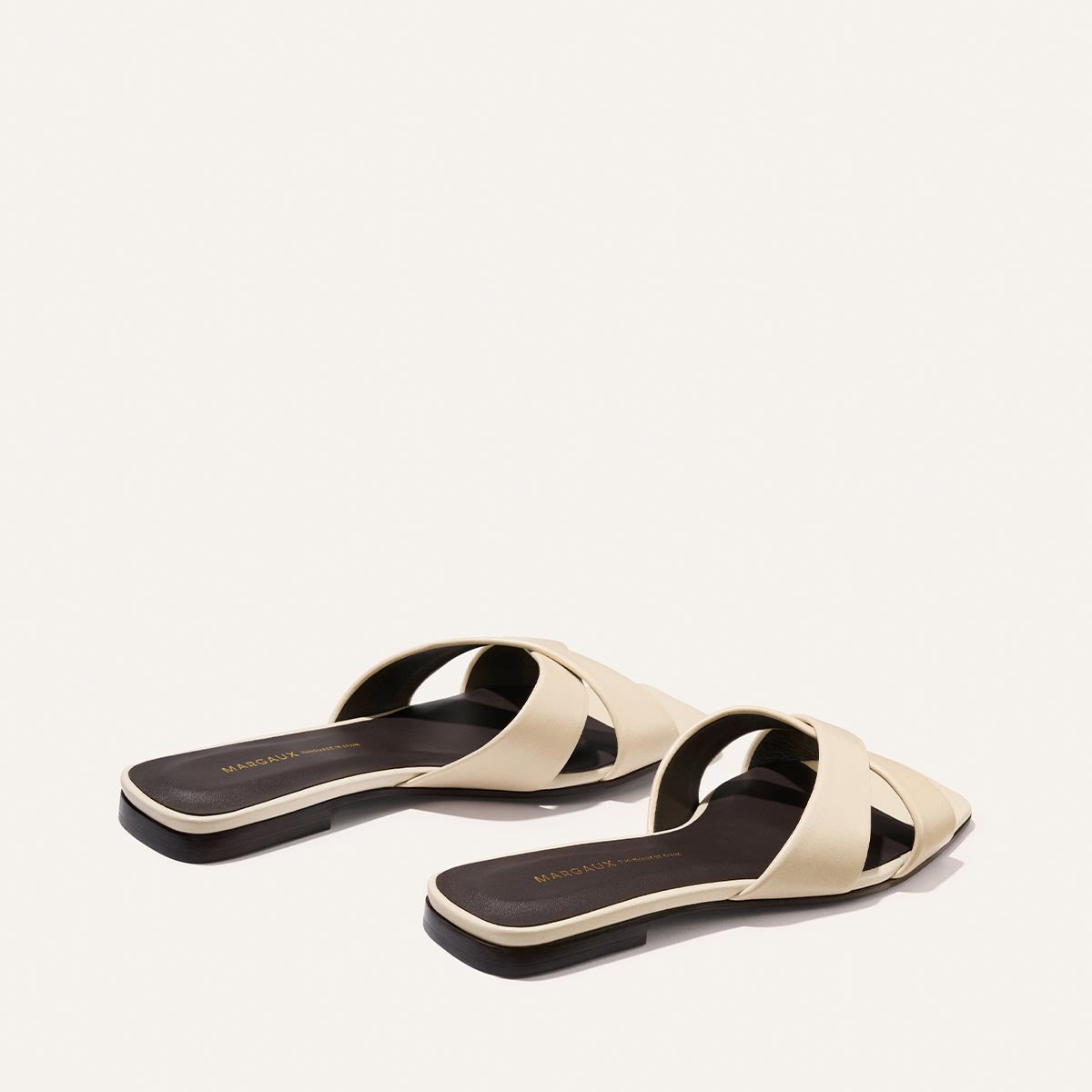 The Selene Flat Sandal - Ivoire Nappa by Margaux features beige slip-on crisscross straps and flat black soles, showcased on a light background.