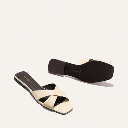 The Selene Flat Sandal - Ivoire Nappa by Margaux: a pair of beige open-toe sandals with crisscross straps and black soles, shown on a white background—one upright, the other flipped to display the sole.