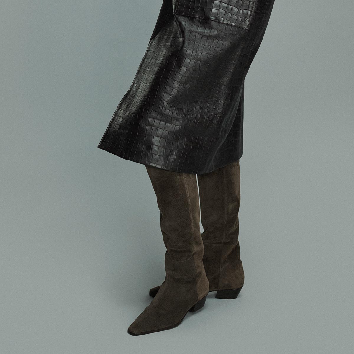 A person wearing a black, textured leather skirt and The Sierra Knee-High Boot in English Brown Suede by Margaux stands against a plain gray background.