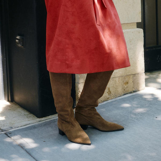 A person wearing a red skirt and Margaux’s The Hayes Knee-High Boot in Driftwood Suede stands on a sunlit sidewalk next to a stone wall and black door.