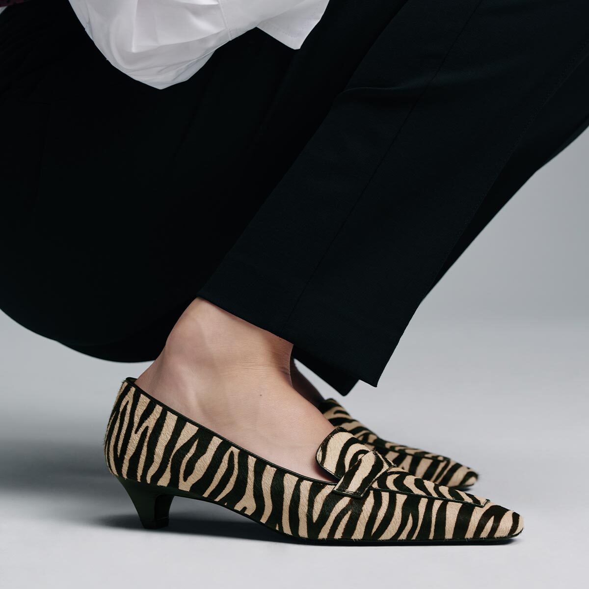 The Sigrid - Tan Zebra Haircalf low-heeled loafers worn with black trousers.