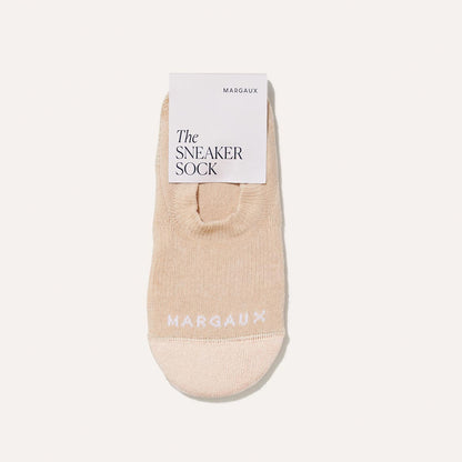 A single beige no-show sock, The Sneaker Sock - Dune by Margaux, features MARGAUX in white near the toe and is displayed flat with a branded card reading Margaux The Sneaker Sock for ultimate on-the-go comfort.