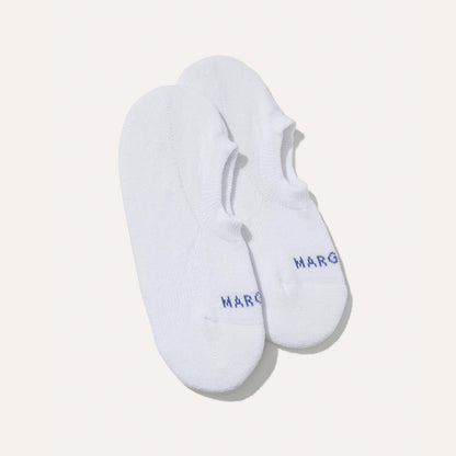 A pair of Margaux’s The Sneaker Sock in white, featuring cushioned no-show design, non-slip grips, and “MARG” embroidered in blue near the toes, shown on a plain light background.