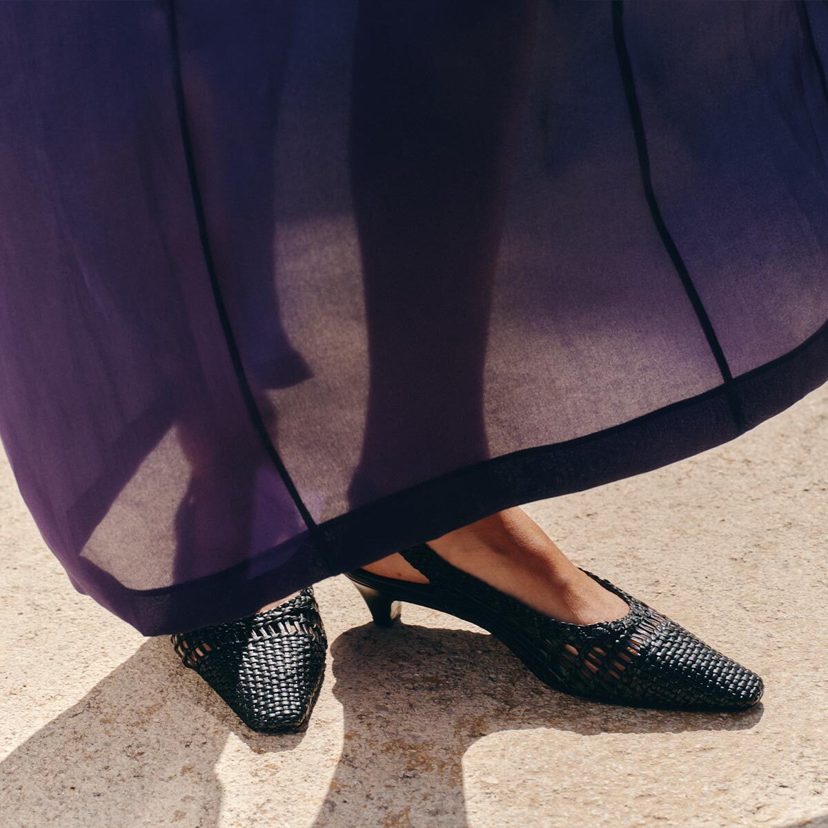 A person wearing a sheer purple skirt and Margaux's The Sofia Sling black woven leather heels with a 1.5" heel stands on a sunlit concrete surface.