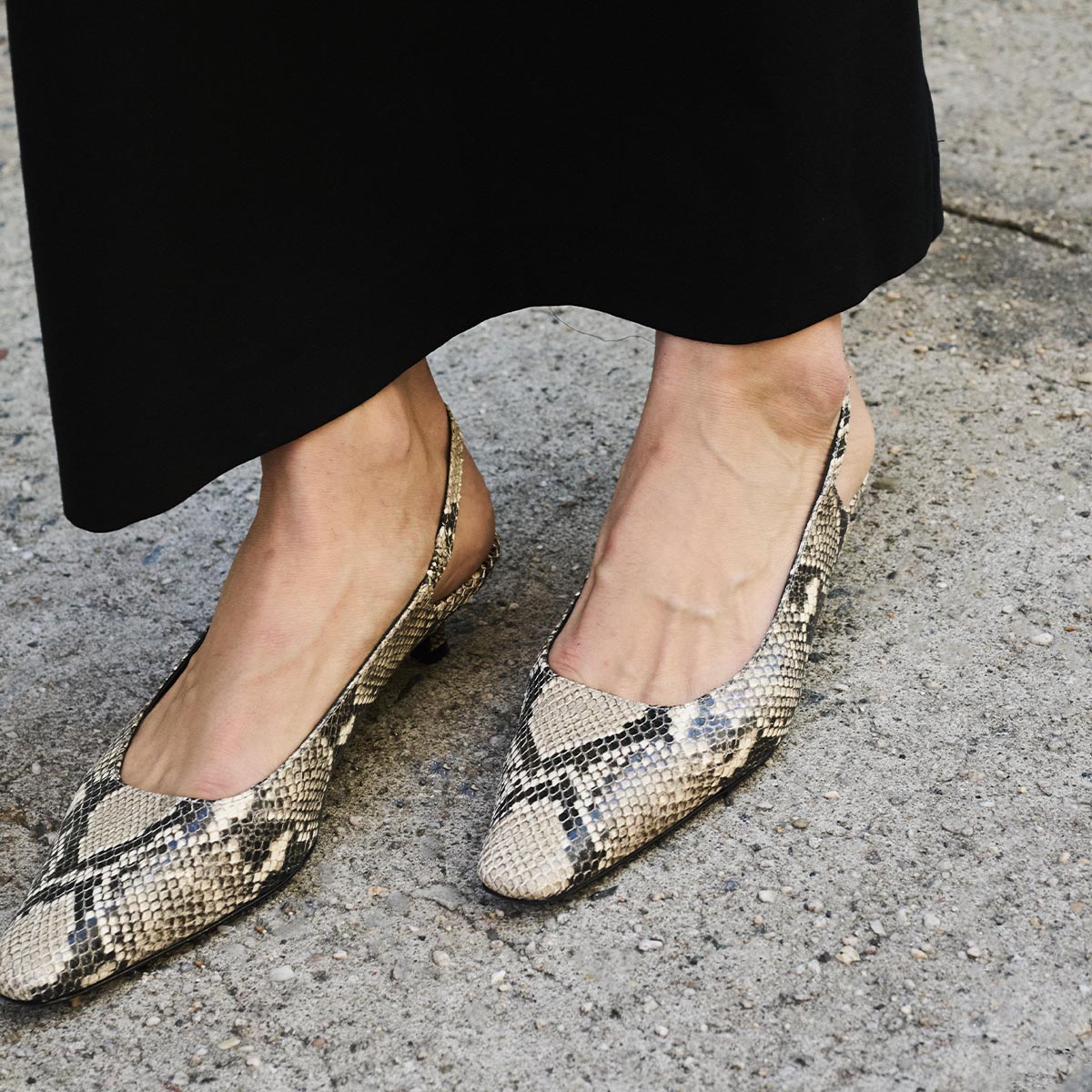 A person wearing a long black skirt and Margauxs The Sofia Sling in Natural Python Embossed stands on a rough concrete surface.