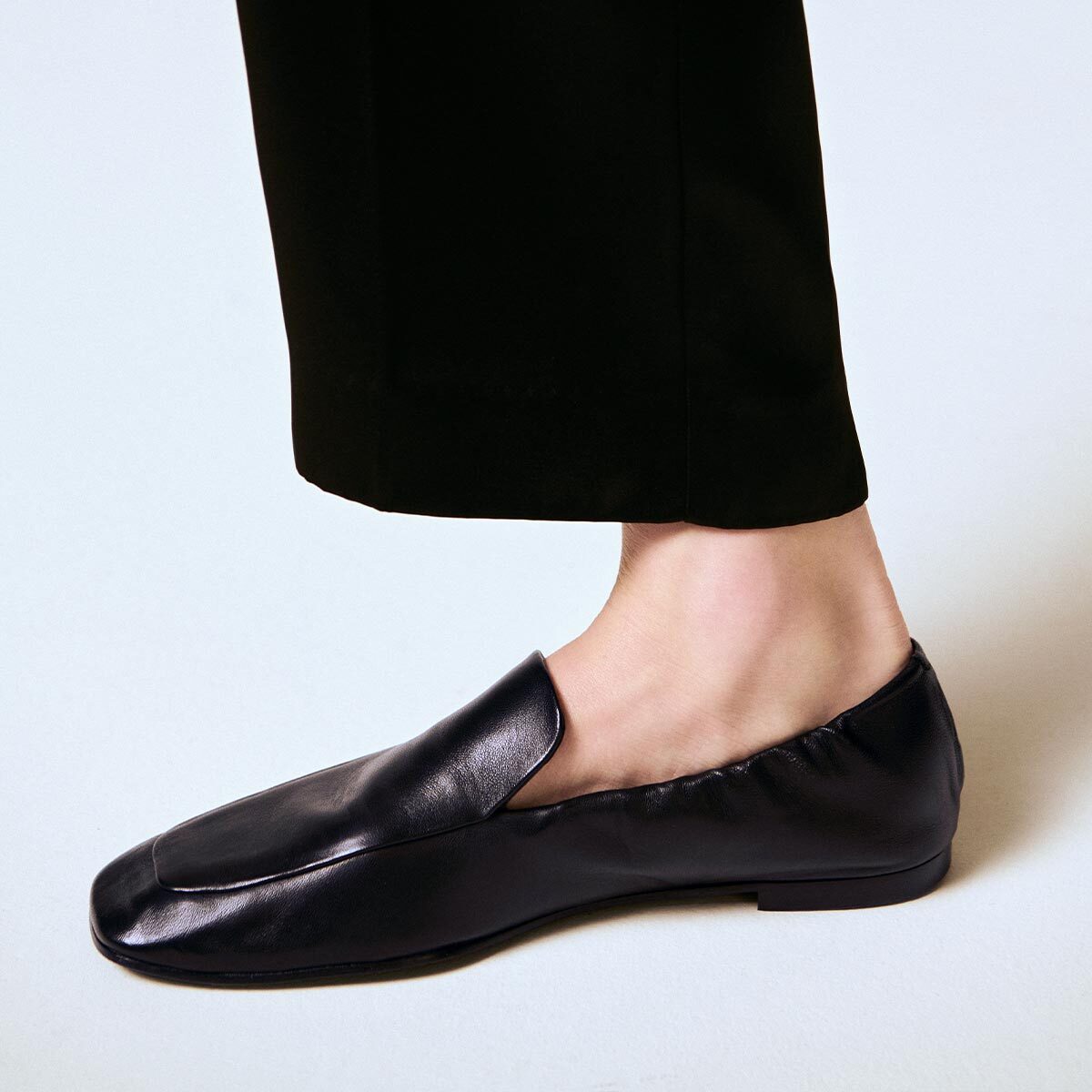 A person wearing black dress pants and Margaux’s The Studio Loafer in Black Nappa stands on a light-colored surface. Only the lower leg and foot are visible.