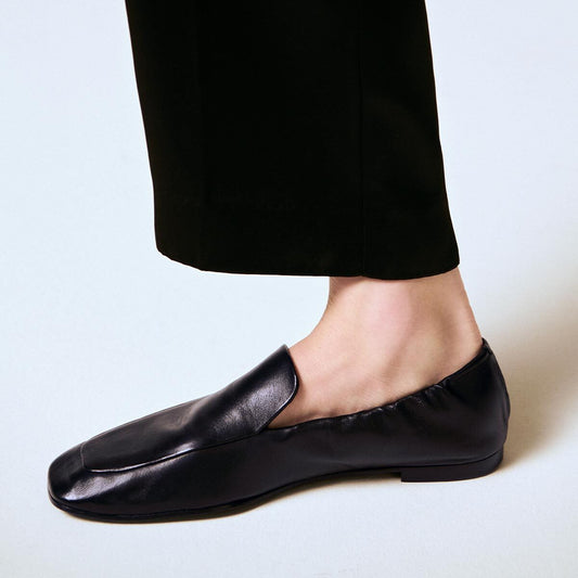 A person wearing black dress pants and Margaux’s The Studio Loafer in Black Nappa stands on a light-colored surface. Only the lower leg and foot are visible.