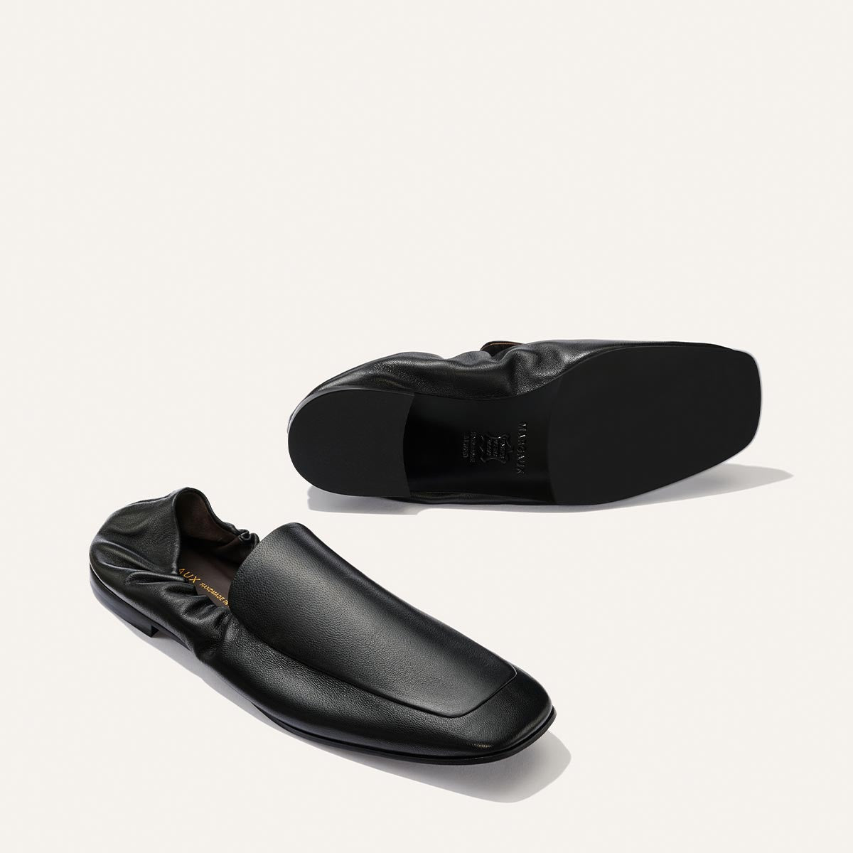 Two Margaux Studio Loafers in Black Nappa are displayed on a plain white background. One shoe is upright, highlighting the top and side view, while the other rests on its side, revealing the sole.