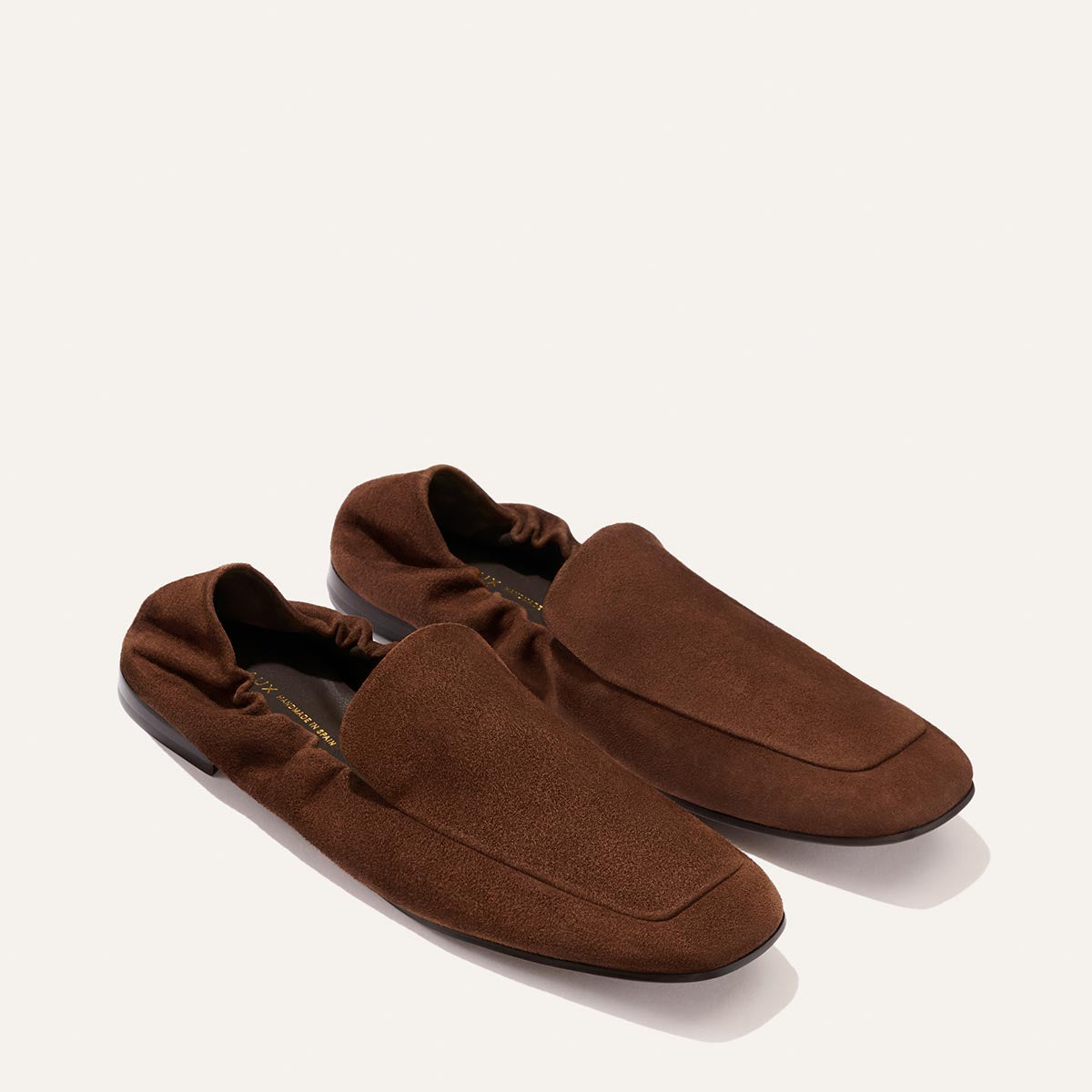 Introducing The Studio Loafer in Ganache Suede by Margaux—a pair of minimalist brown suede loafers, designed with elasticized backs and subtly squared toes, elegantly showcased against a simple light background.
