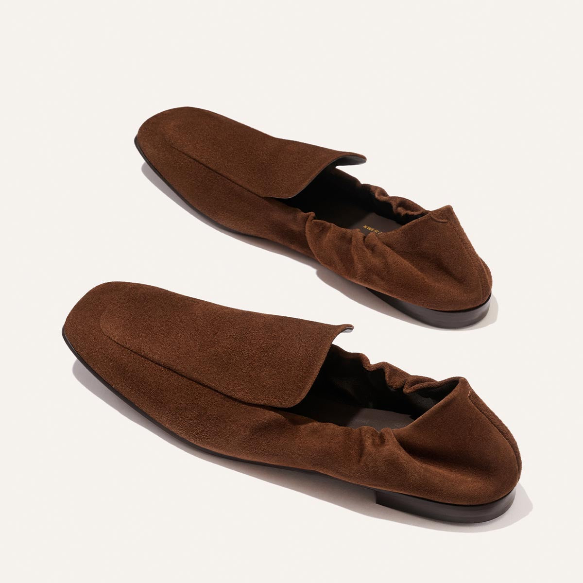 The Studio Loafer - Ganache Suede by Margaux is a pair of brown suede slip-on loafers with an elasticized back, displayed against a light, neutral background. Featuring a minimalist design with flat soles and no visible laces or embellishments, these shoes offer understated elegance.