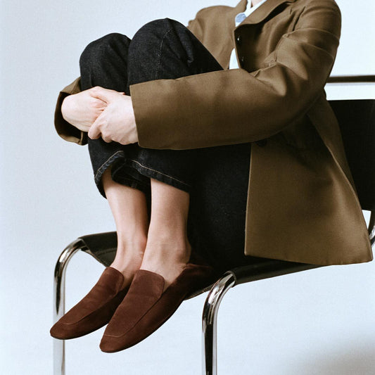 A person sits on a chair with their knees up, arms wrapped around their legs, wearing a brown blazer, dark jeans, and the Margaux Studio Loafer in Ganache Suede—a brown loafer featuring an elongated square toe.